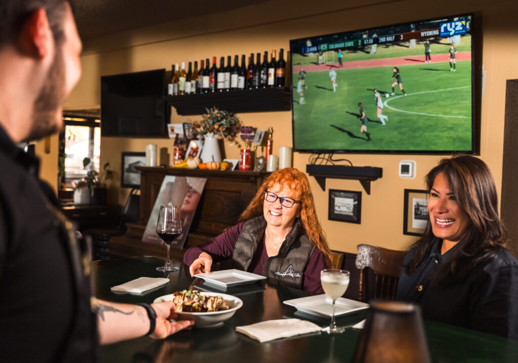 Two women enjoying drinks at bar with TV sports