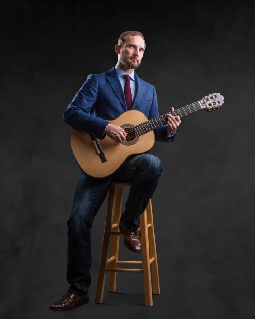 vjh Man playing acoustic guitar on a stool.