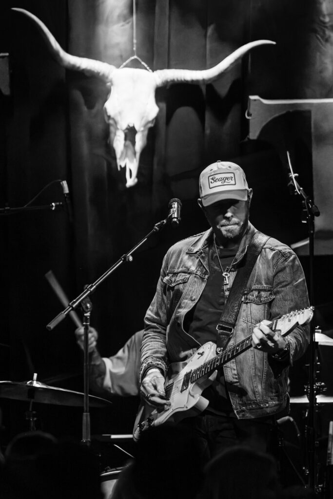 Musician on stage with guitar, bull skull backdrop.