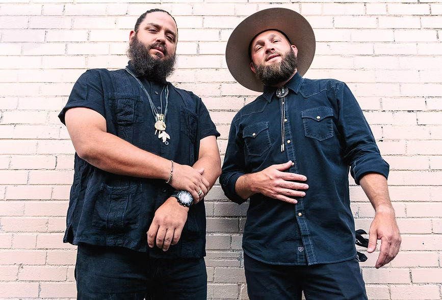 Two bearded men in dark outfits against brick wall