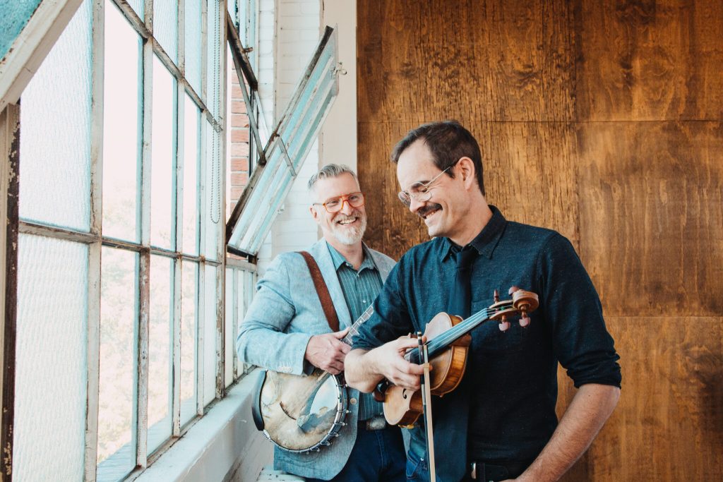 Two smiling musicians holding banjo and violin by window