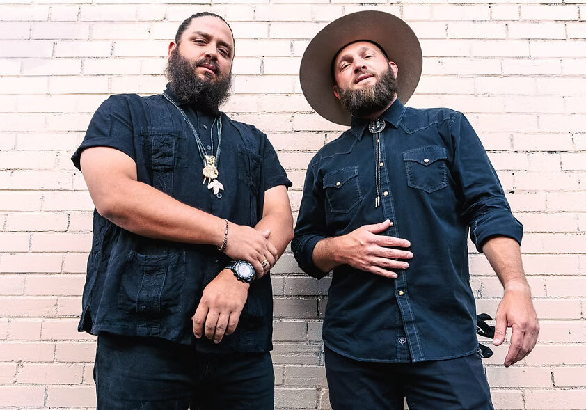 Two bearded men in dark outfits against brick wall