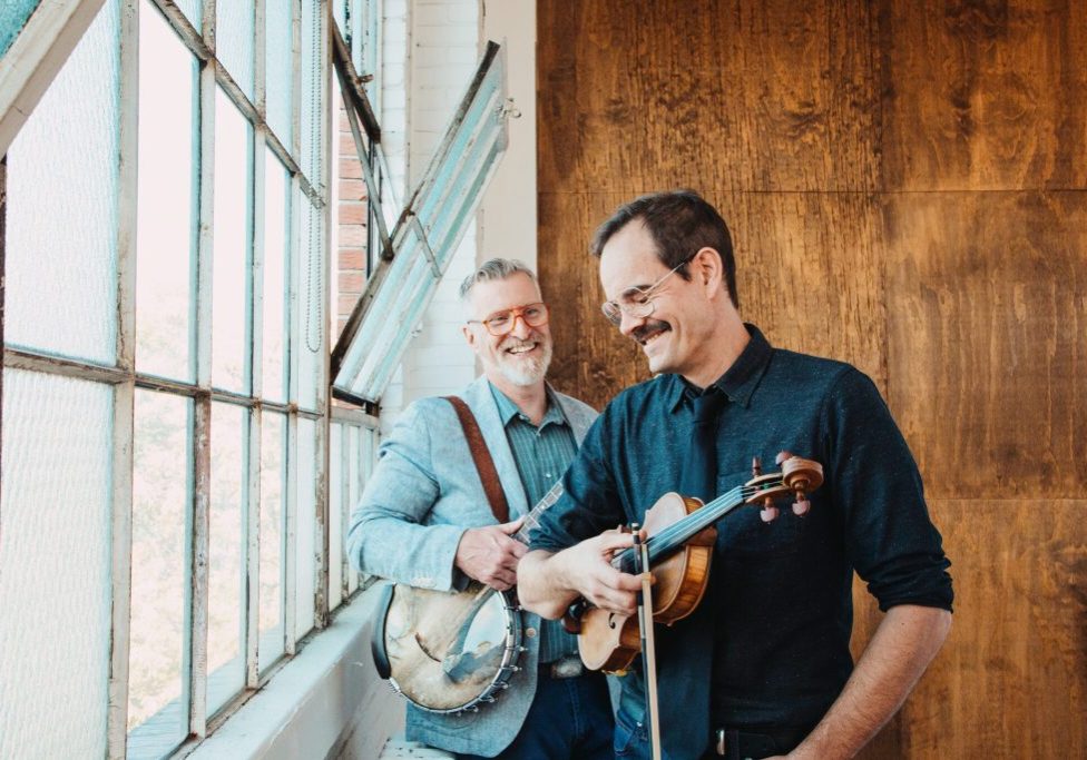 Two smiling musicians holding banjo and violin by window